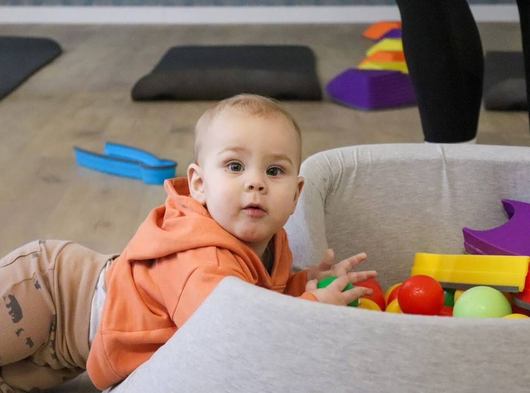 Kleinkind in orangefarbenem Kapuzenpulli spielt im Kursraum mit bunten Bällen