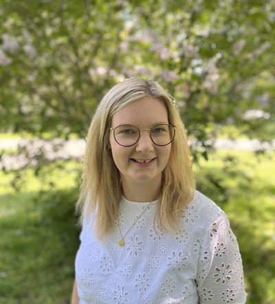 Birte Görmar mit Brille und weißer Spitzenbluse vor blühenden Sträuchern