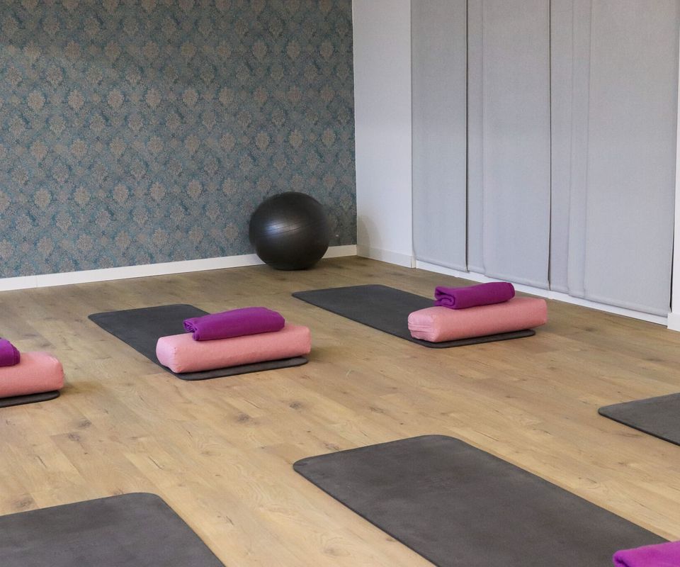 Kursraum mit hellem Holzboden, ausgelegten Gymnastikmatten, rosa Nackenrollen und violetten Handtüchern, hinten ein schwarzer Gymnastikball vor gemusterter Tapete